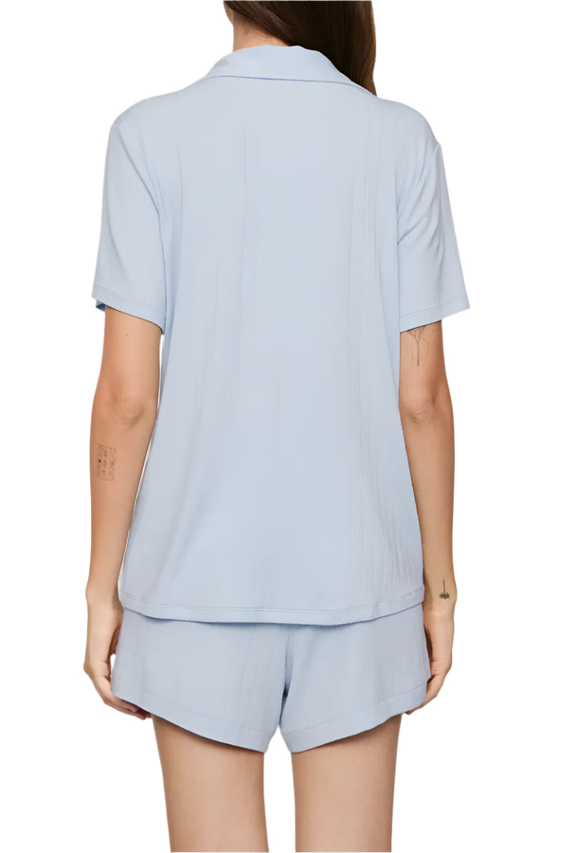 A person stands with their back to the camera wearing the Eberjey Gisele Rib Relaxed Short PJ Set, featuring a light blue button-down pajama top and matching shorts with an elastic waistband.