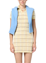 A person in a yellow Plaid Polo Dress by Guest in Residence with short sleeves and a blue sweater over their shoulders, accessorized with a beaded necklace, stands against a white background.