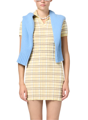 A person in a yellow Plaid Polo Dress by Guest in Residence with short sleeves and a blue sweater over their shoulders, accessorized with a beaded necklace, stands against a white background.