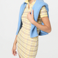 Wearing the Guest in Residence Plaid Polo Dress, a yellow silk linen blend with short sleeves, paired with a blue sweater over the shoulders, this person stands against a plain light background.