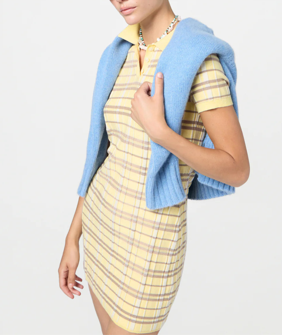 Wearing the Guest in Residence Plaid Polo Dress, a yellow silk linen blend with short sleeves, paired with a blue sweater over the shoulders, this person stands against a plain light background.