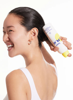A woman with wet hair in a white tank top smiles as she applies Supergoop Play Antioxidant Mist SPF 50 with Vitamin C (3 fl oz) from a yellow and white can to the back of her head.