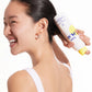 A woman in a white tank top applies Supergoop Play Antioxidant Mist SPF 50 with Vitamin C (6 fl oz) to her neck and hairline, smiling sideways against a plain background, capturing the glow of sun-protected skin.