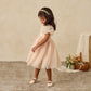 A young girl wears the Noralee Baby Girls' and Girls' Dorothy Dress in light pink with puff sleeves and an embroidered bodice, standing on a textured rug. Behind her is a basket filled with flowers and fabric.
