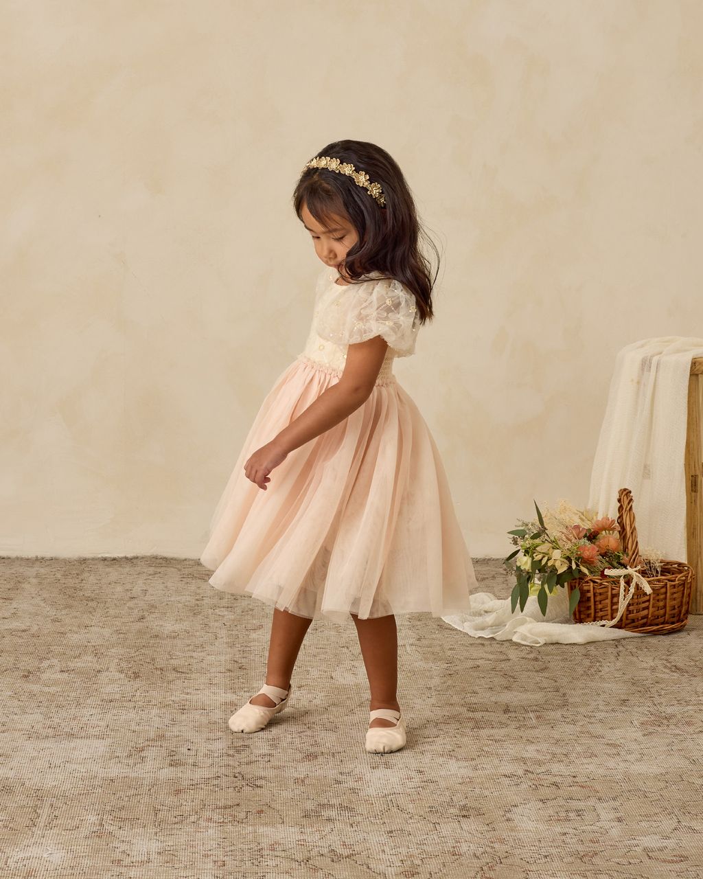 A young girl wears the Noralee Baby Girls' and Girls' Dorothy Dress in light pink with puff sleeves and an embroidered bodice, standing on a textured rug. Behind her is a basket filled with flowers and fabric.