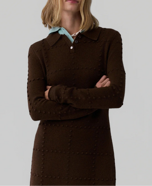 A person with shoulder-length blonde hair wears the Popcorn Polo Dress by Guest in Residence—a brown women’s sweater dress featuring a collared neckline and buttons—standing with arms crossed.