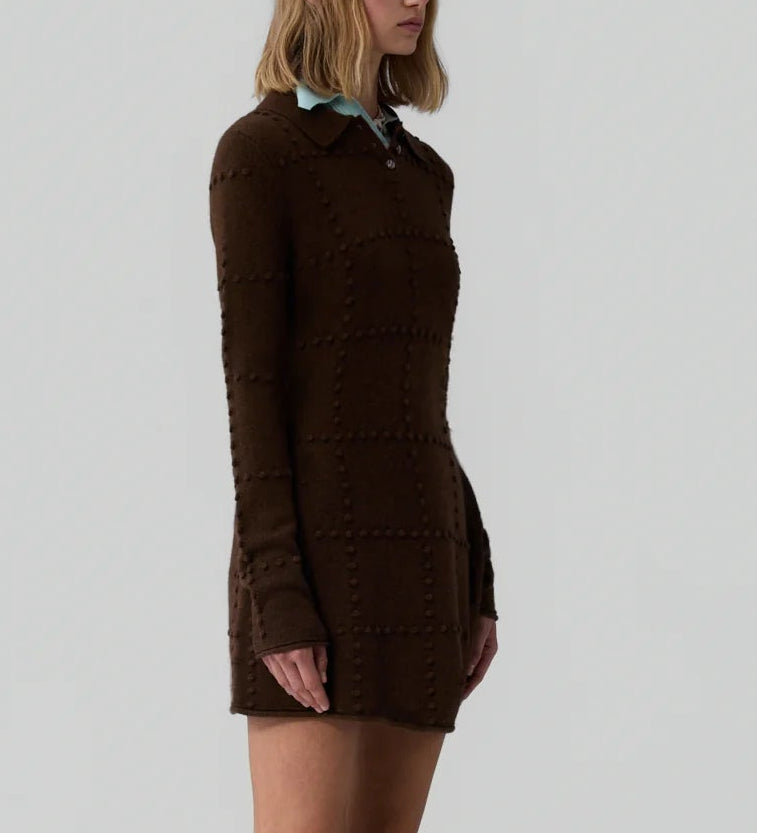 A woman with shoulder-length blonde hair wears the Popcorn Polo Dress by Guest in Residence—a long-sleeved, brown textured sweater dress with a collar—standing against a plain light background.