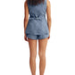 A woman stands facing away in POSSE's Thea Short—a blue, high-rise waist short—paired with a matching sleeveless belted vest and flat black shoes, set against a white background.