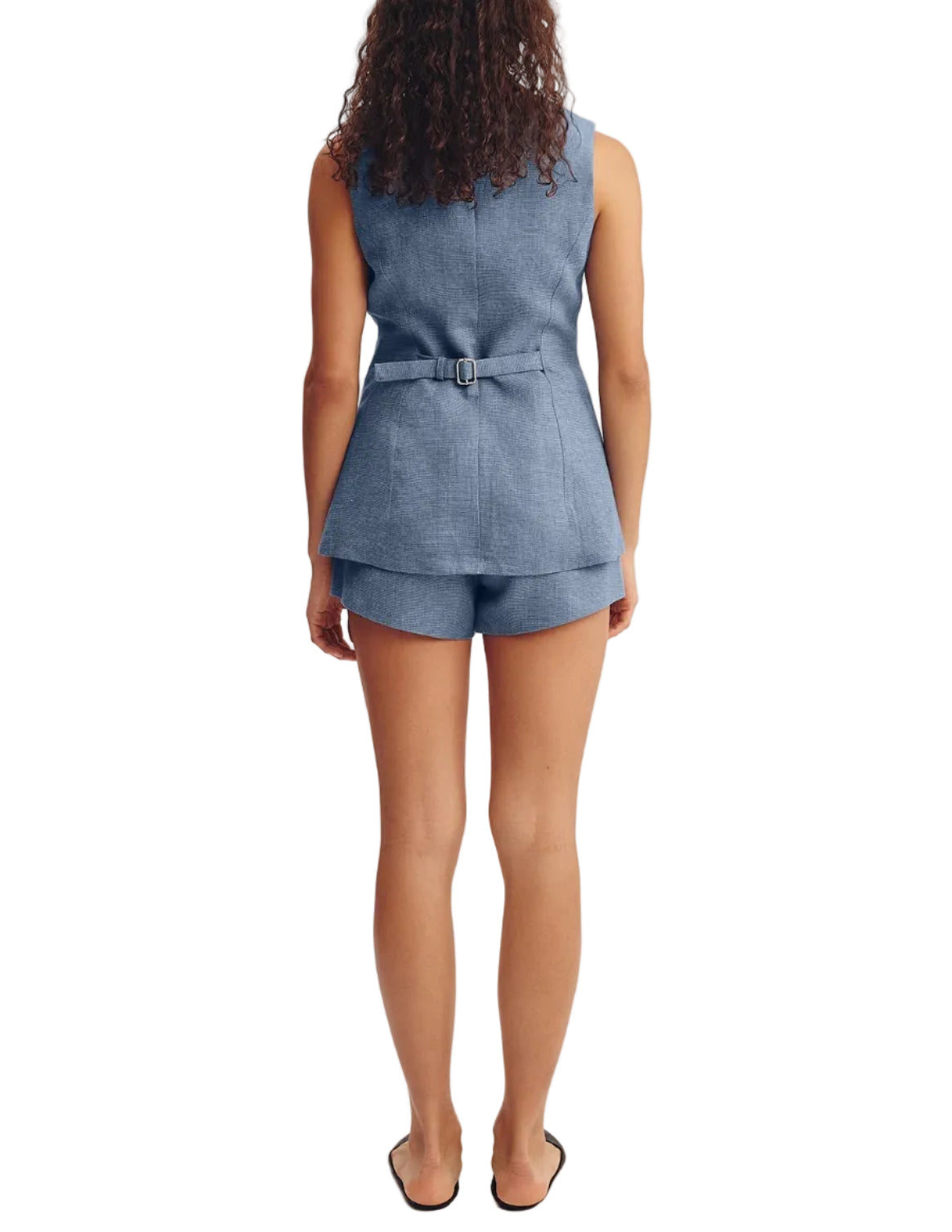 A woman stands facing away in POSSE's Thea Short—a blue, high-rise waist short—paired with a matching sleeveless belted vest and flat black shoes, set against a white background.