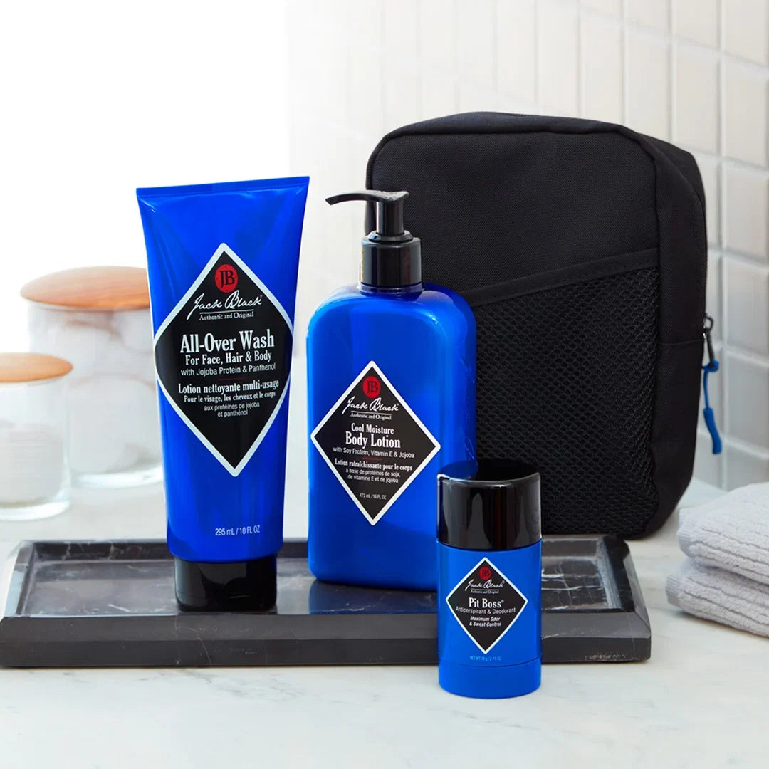 The Jack Black Clean and Cool Body Basics Set, featuring three blue grooming essentials for daily body care, is displayed on a tray along with a black toiletry bag and a folded white towel on a bathroom countertop.