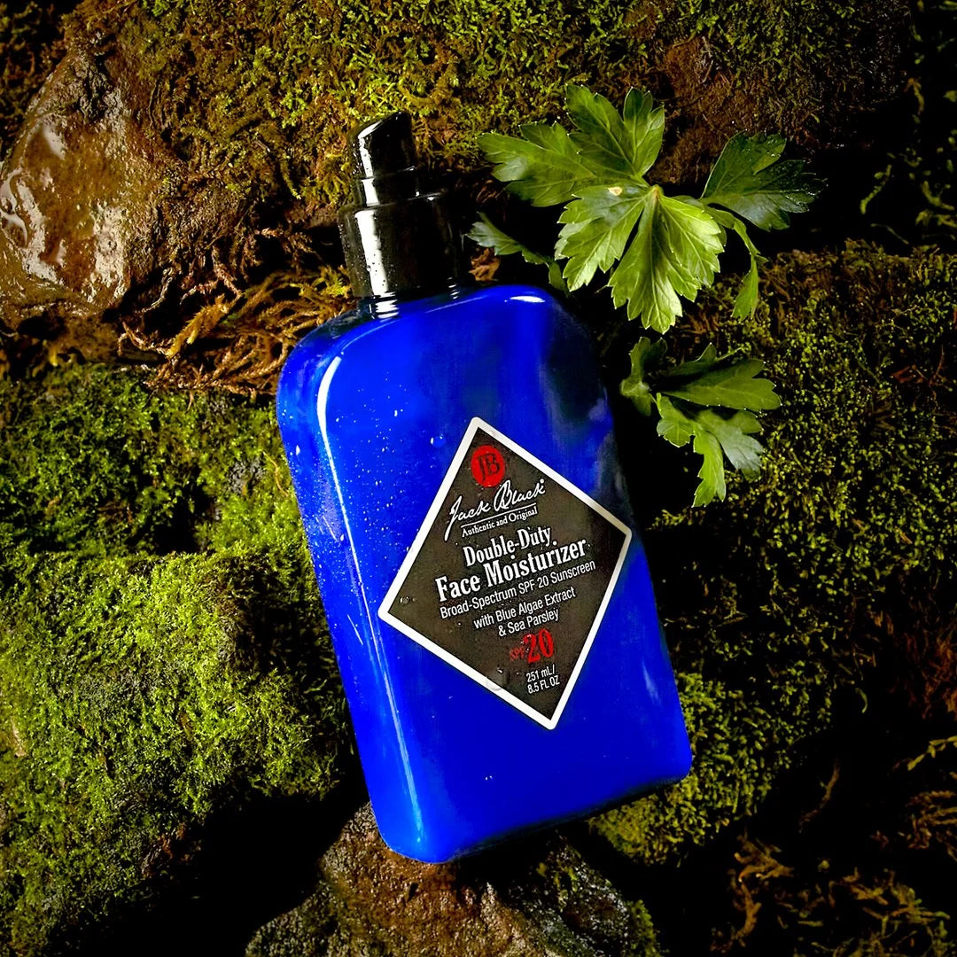 A blue bottle of Jack Black Double-Duty Face Moisturizer sits on green moss beside fresh parsley leaves.