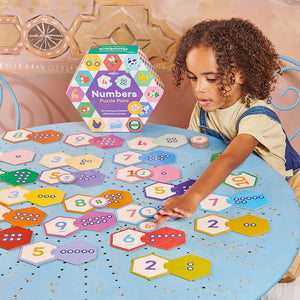A young child practices counting and math by matching numbered hexagon pieces with dot groups from the Eeboo Numbers Hexagon Puzzle Pairs set at a round blue table.