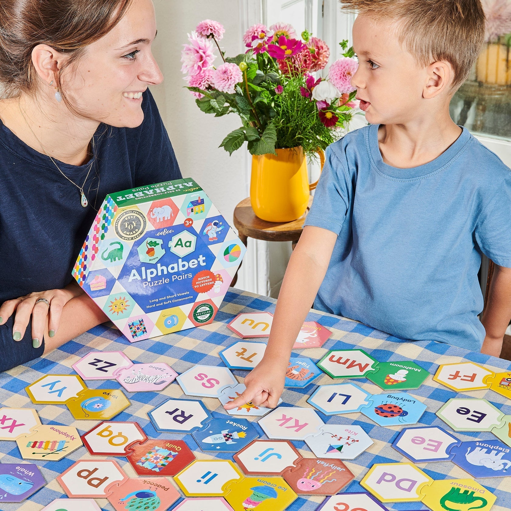 A woman and a child enhance their phonological awareness by playing with the Eeboo Alphabet Hexagon Pairing Puzzles on a checkered table. In the background, a vase with pink flowers blooms gently, adding a touch of warmth to this moment of nurturing pre-literacy skills.