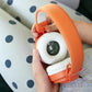 Person holding Yoto, Headphones Orange by Yoto with a cartoon design inside the ear cup, against a background with polka dots. These headphones feature kid-safe volume limiting and an adjustable headband for a comfortable fit.