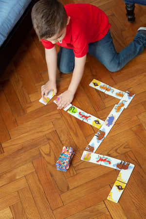A boy in a red shirt kneels on a wooden floor, arranging Eeboo’s Animal Parade Head to Toe Dominoes in a winding pattern, making this classic game by Eeboo into a fun travel adventure.