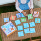 A child plays Eeboo's Pre-school Nice Animals Memory Game, matching animal cards on a wicker table outdoors, while an adult sits nearby.