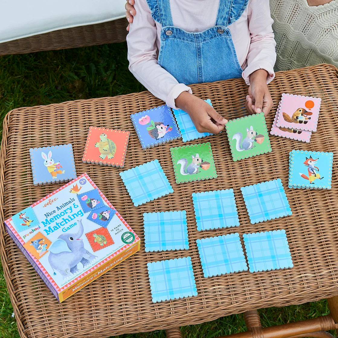 A child plays Eeboo's Pre-school Nice Animals Memory Game, matching animal cards on a wicker table outdoors, while an adult sits nearby.