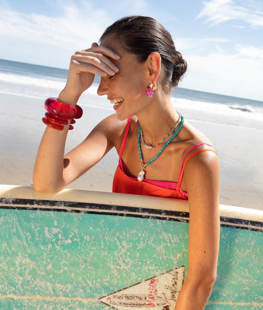 A person in a red dress holds a surfboard on the beach, adorned with colorful jewelry and Lizzie Fortunato Organic Hoops, smiling under the sunny sky.