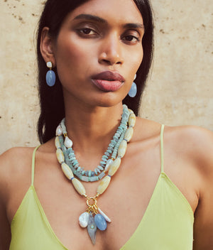 A woman in a light yellow spaghetti strap top wears layered beaded necklaces with mother-of-pearl accents and Lizzie Fortunato’s Big Sur Earrings, standing against a textured beige background.