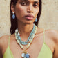Wearing a light green top, a Lizzie Fortunato Marilla Necklace of blue and white beads with gold accents, and matching blue drop earrings, a person poses against a neutral background.