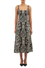 A woman wears the Ulla Johnson Natali Tank Dress—a sleeveless black poplin dress with a white floral leaf print and square neckline—paired with black pointed ankle-strap shoes.