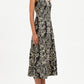 A woman stands against a white background wearing the Ulla Johnson Natali Tank Dress, a sleeveless black midi dress with a white floral leaf print, paired with black pointed-toe heels.