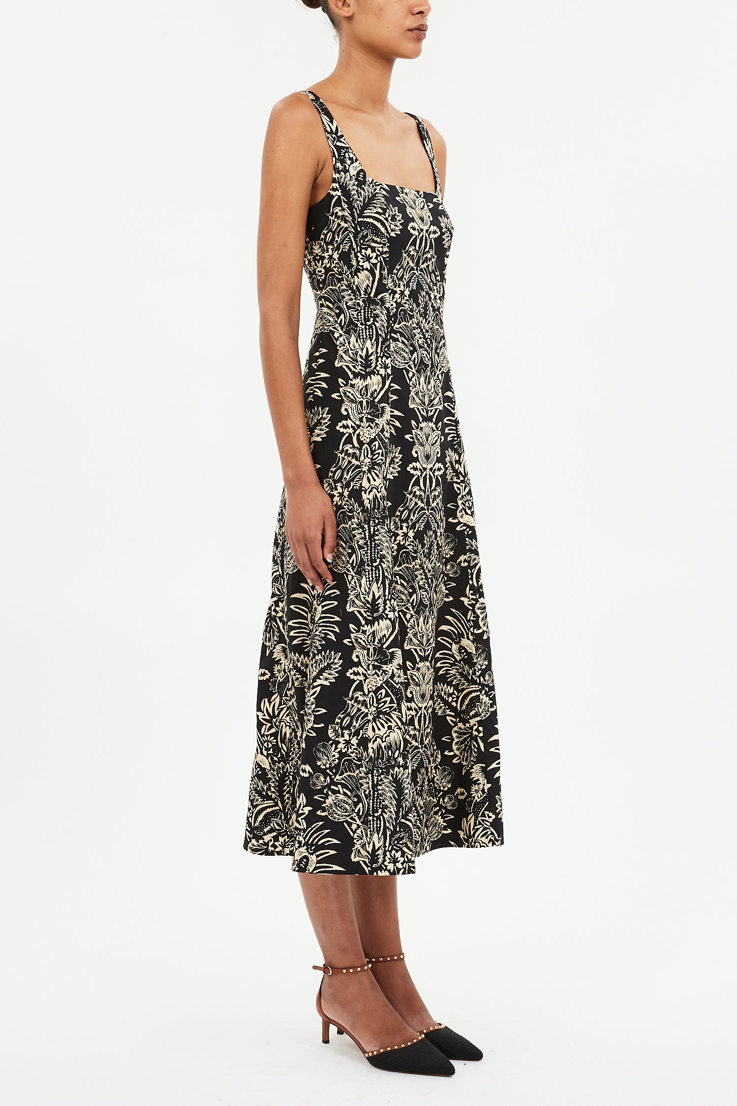 A woman stands against a white background wearing the Ulla Johnson Natali Tank Dress, a sleeveless black midi dress with a white floral leaf print, paired with black pointed-toe heels.