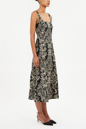 A woman stands against a white background wearing the Ulla Johnson Natali Tank Dress, a sleeveless black midi dress with a white floral leaf print, paired with black pointed-toe heels.