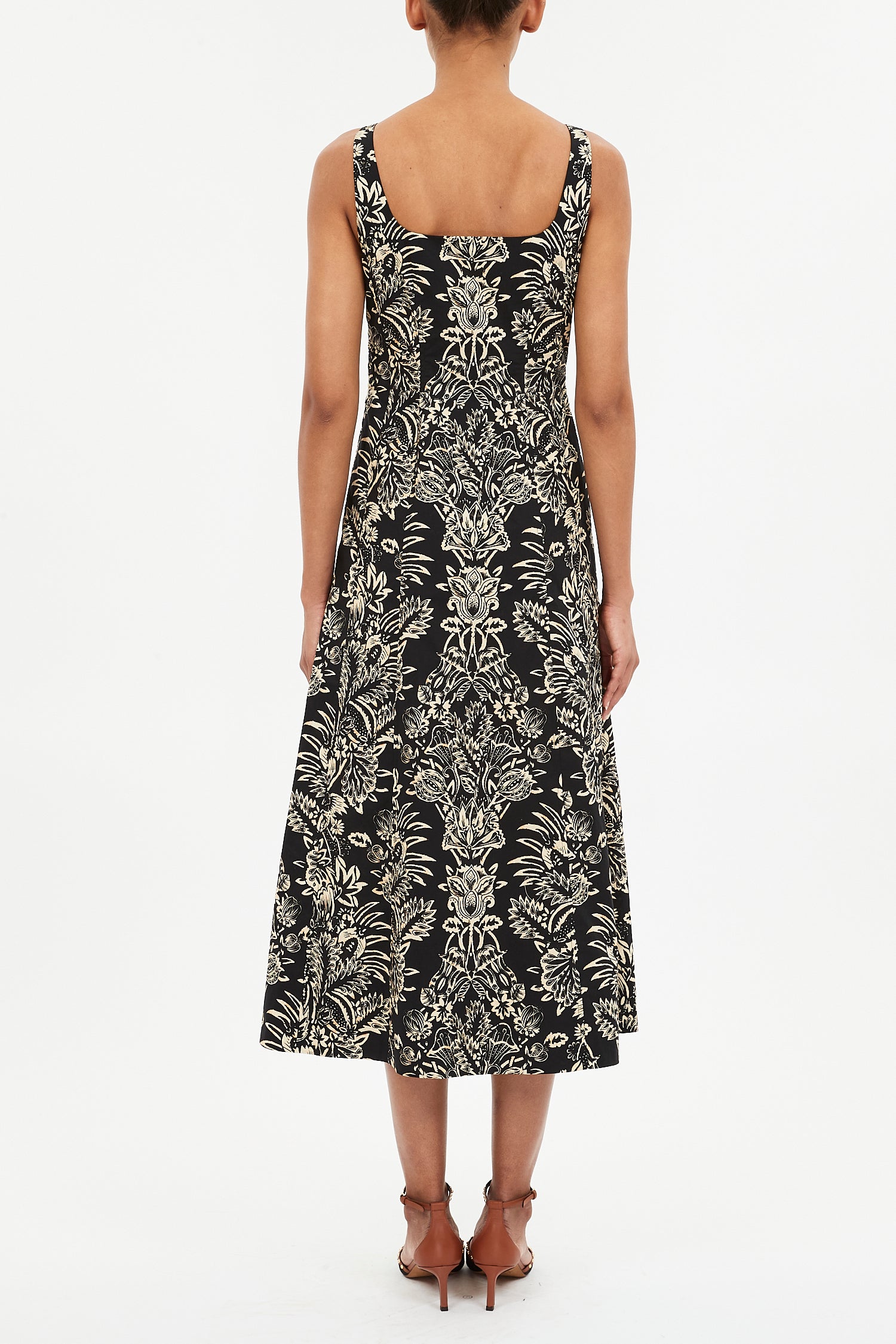 A person stands facing away in the Ulla Johnson Natali Tank Dress—sleeveless, black, with a beige floral leaf print—paired with brown heeled sandals against a plain white background.