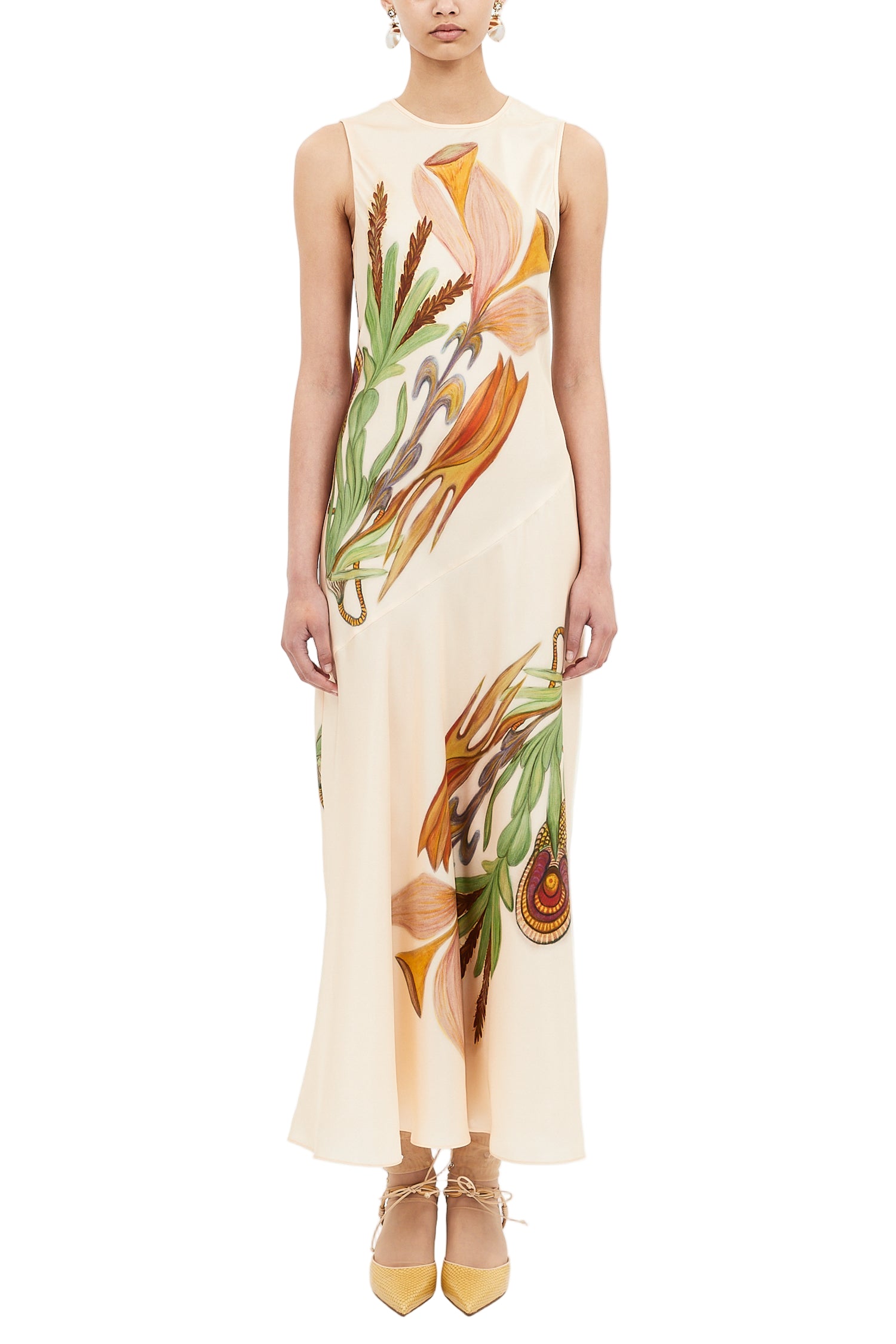 A person wearing the Ulla Johnson Gaea Dress, a cream sleeveless midi with bold florals and an ankle-length bias cut, styled with tan pointed-toe shoes.