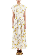 A person wearing the Ulla Johnson Lausanne Dress, a white, ankle-length floral print with yellow and blue flowers and cap sleeves, paired with brown flat shoes.