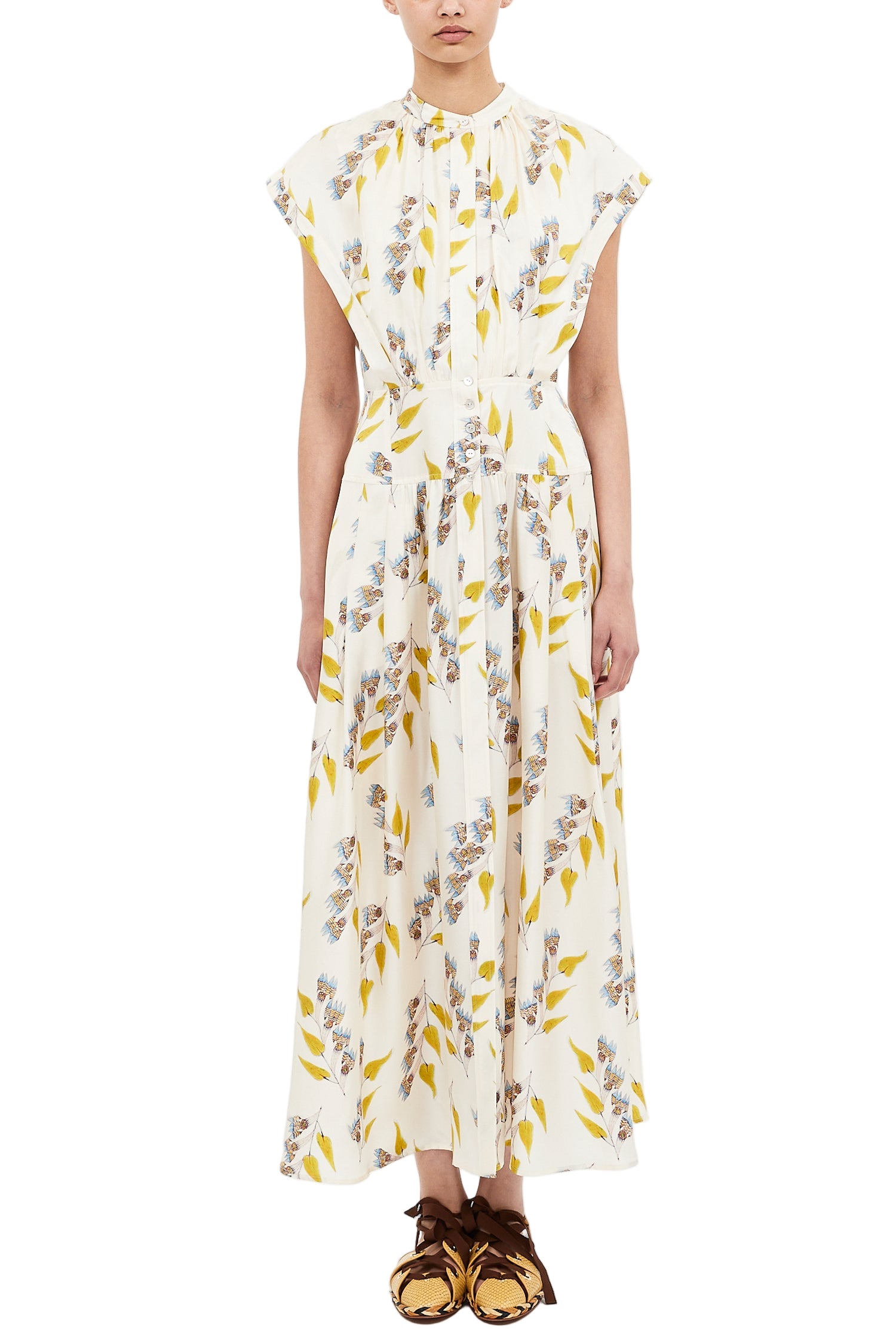 A person wearing the Ulla Johnson Lausanne Dress, a white, ankle-length floral print with yellow and blue flowers and cap sleeves, paired with brown flat shoes.
