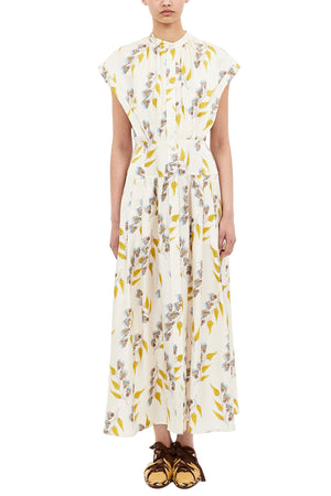 A person wearing the Ulla Johnson Lausanne Dress, a white, ankle-length floral print with yellow and blue flowers and cap sleeves, paired with brown flat shoes.