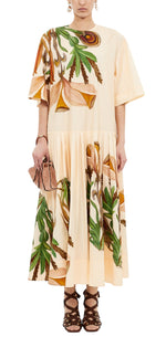 A person wears the Ulla Johnson Aniline Drop Waist Cotton Dress in light tan with green and brown botanical prints, paired with a beige clutch and brown lace-up sandals.