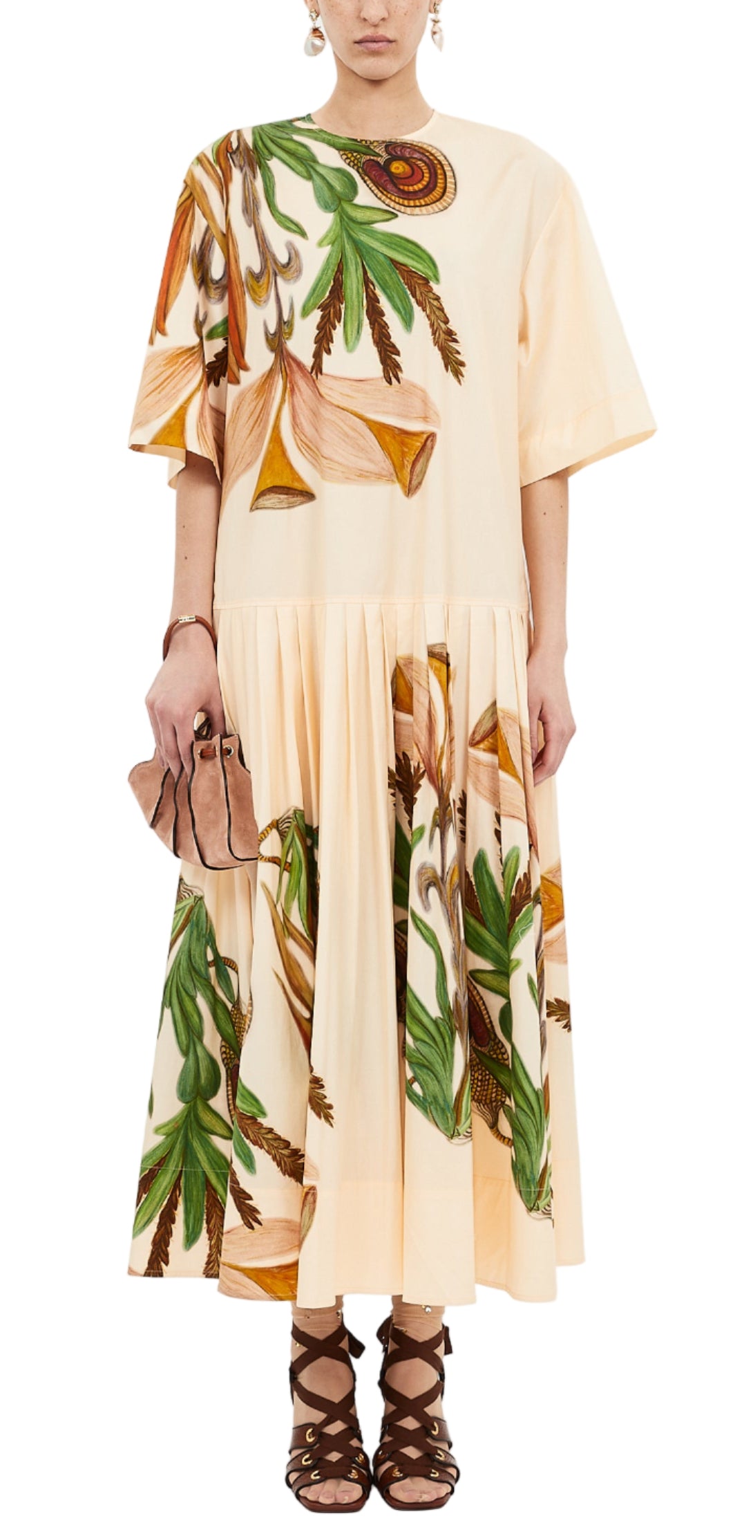 A person wears the Ulla Johnson Aniline Drop Waist Cotton Dress in light tan with green and brown botanical prints, paired with a beige clutch and brown lace-up sandals.