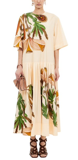 A person wears the Ulla Johnson Aniline Drop Waist Cotton Dress in light tan with green and brown botanical prints, paired with a beige clutch and brown lace-up sandals.