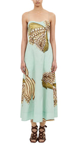 A woman wears the Ulla Johnson Vesela Cami Midi Dress — a light blue dress with thin straps and an abstract beige and brown floral print — paired with lace-up sandals, standing against a white background.