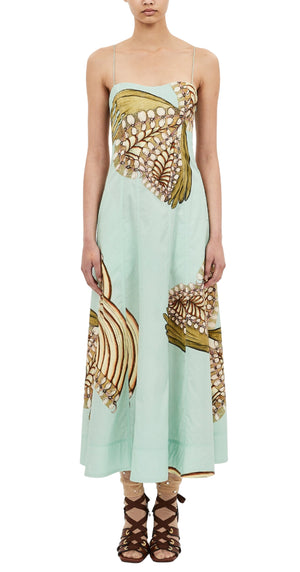 A woman wears the Ulla Johnson Vesela Cami Midi Dress — a light blue dress with thin straps and an abstract beige and brown floral print — paired with lace-up sandals, standing against a white background.