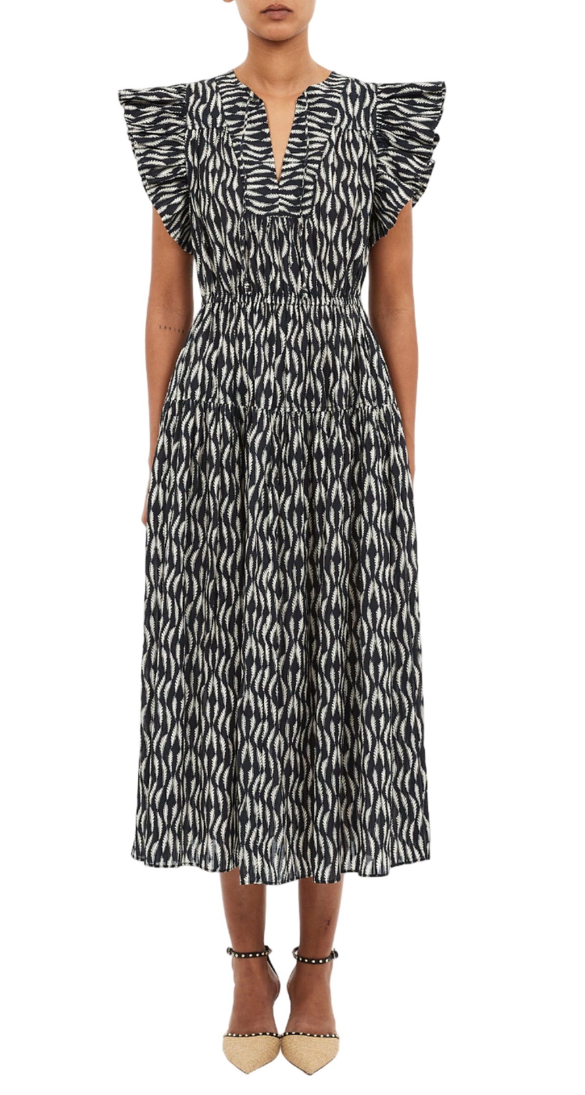 A woman in the Ulla Johnson Kairi Dress—a black and white patterned, ankle-length dress with ruffled short sleeves and V-neck—paired with beige ankle-strap shoes.