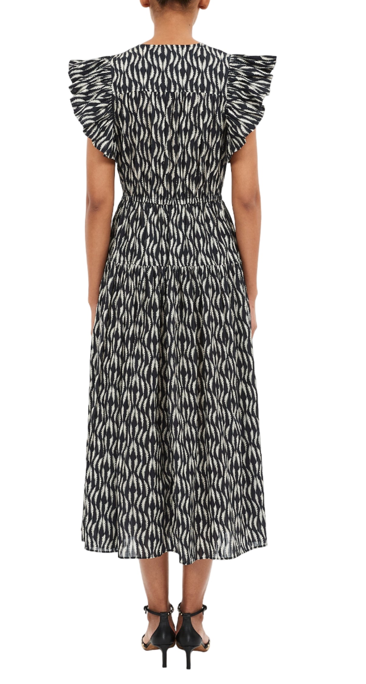A person stands facing away, wearing the Ulla Johnson Kairi Dress—black and white patterned with ruffled sleeves—paired with black strappy sandals.