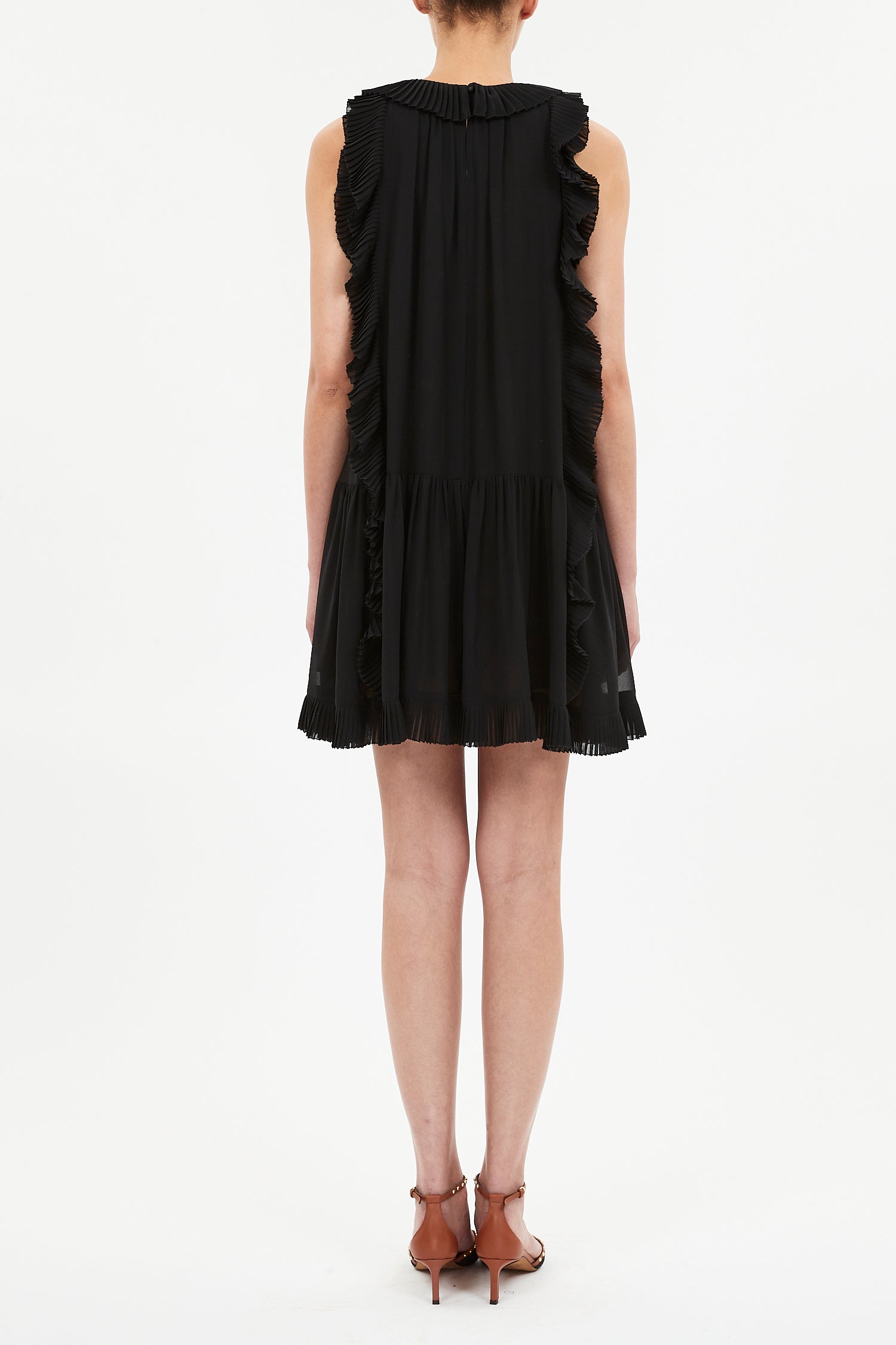 A person in the Ulla Johnson Signe Mini Dress, featuring cascading ruffles and sleeveless silk design, paired with brown heeled sandals, stands facing away from the camera against a plain white background.