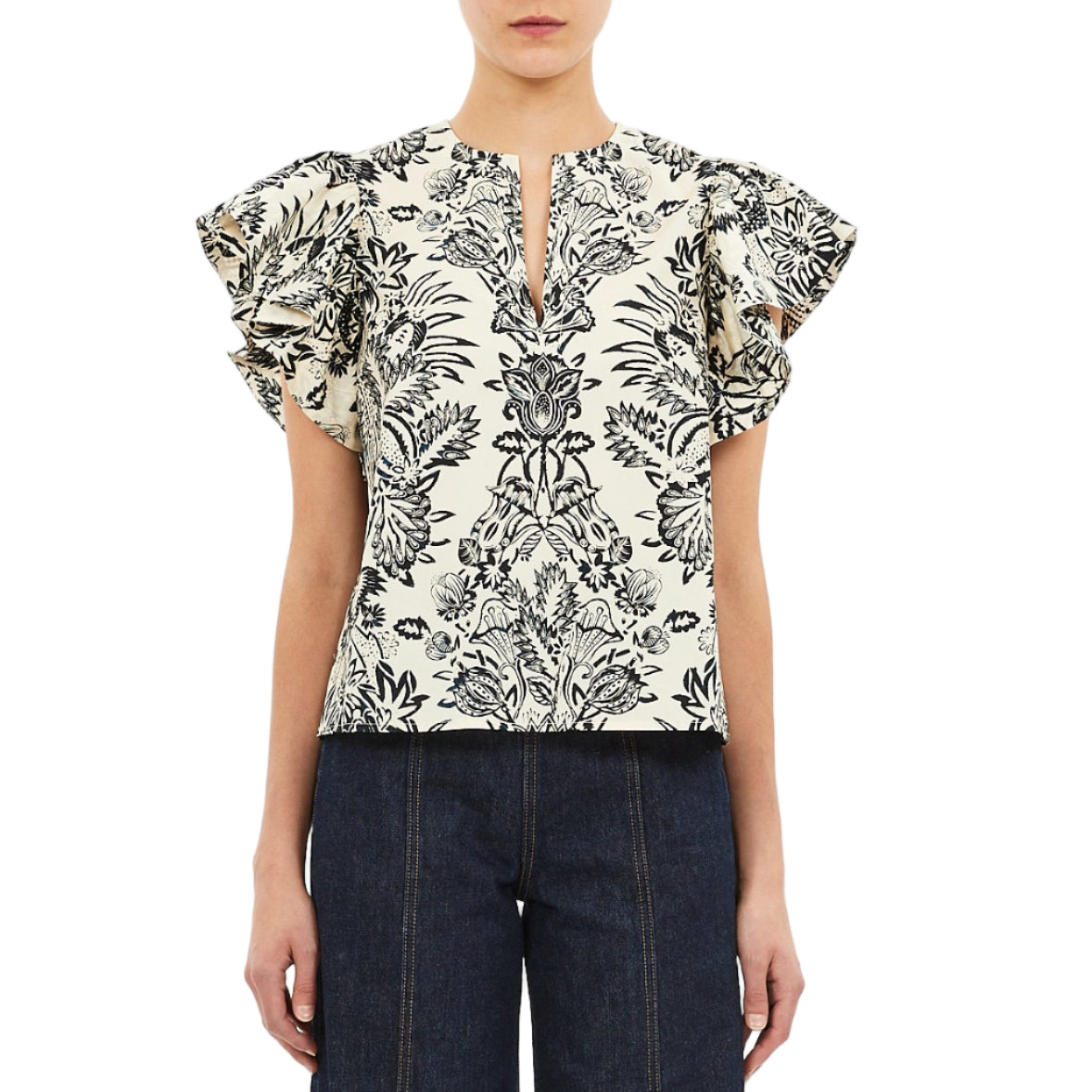 A woman wears the Ulla Johnson Severine Top, a black botanical print flutter sleeve blouse in white cotton poplin, paired with dark denim pants and posed against a white backdrop.