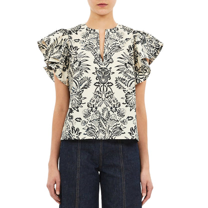 A woman wears the Ulla Johnson Severine Top, a black botanical print flutter sleeve blouse in white cotton poplin, paired with dark denim pants and posed against a white backdrop.