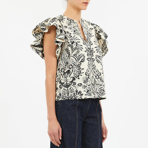 A person wearing the Ulla Johnson Severine Top, a black and white floral V-neck blouse with flutter sleeves, pairs it with dark denim jeans and stands against a plain background.