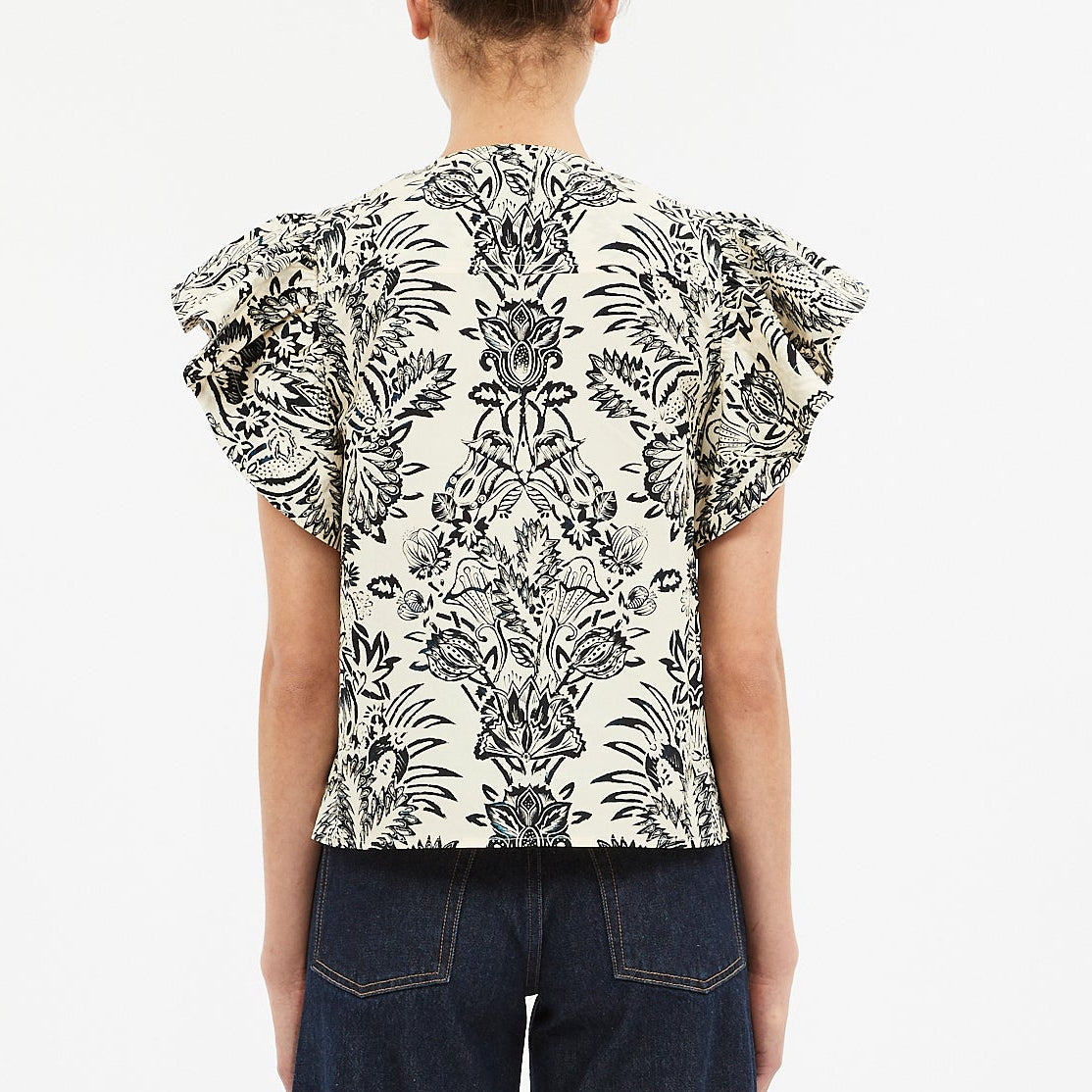 A person is seen from the back wearing the Ulla Johnson Severine Top, a black and white botanical print blouse with ruffled sleeves, paired with dark jeans.