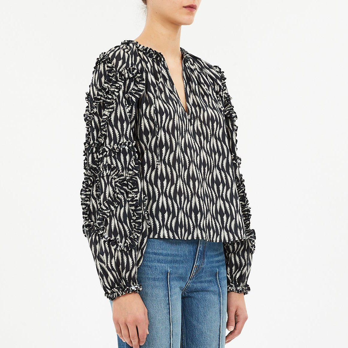 A person wearing the Ulla Johnson Azrou Blouse, featuring a patterned design and ruffled details, paired with blue jeans, shown from the waist up against a plain background.