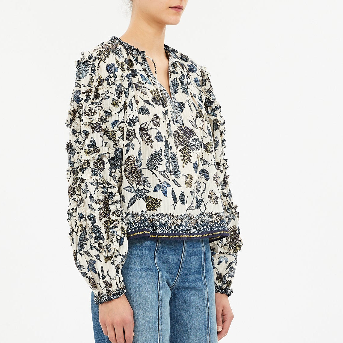 A person is shown from shoulders to hips wearing the Ulla Johnson Azrou Blouse, a long-sleeve, floral-patterned top with ruffled details, paired with blue jeans.