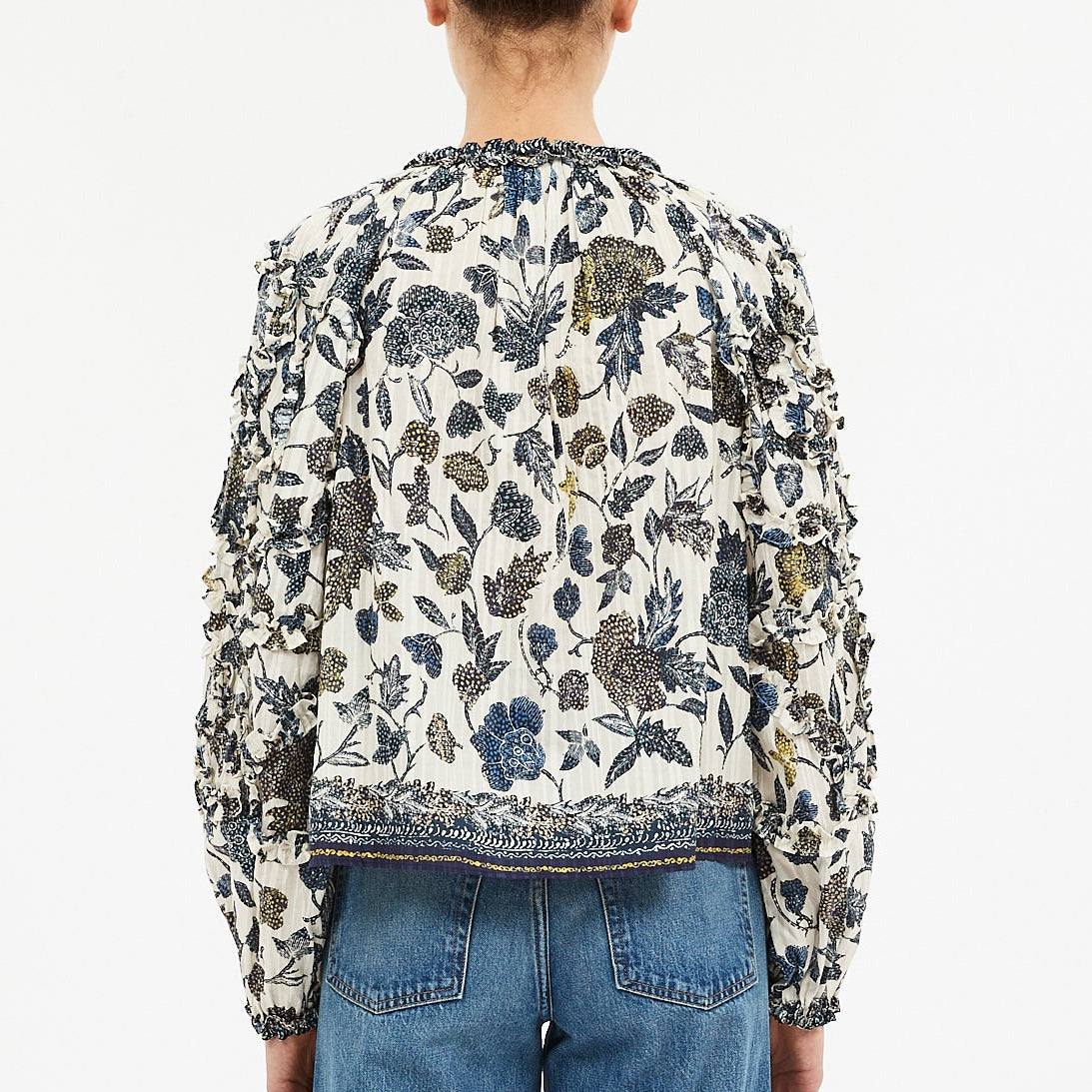 A person is seen from the back wearing the Ulla Johnson Azrou Blouse, a long-sleeved, floral-patterned top with ruffled arm details, paired with blue jeans.
