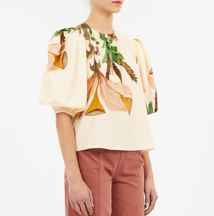 Someone wearing the Ulla Johnson Ravena Puff Sleeve Top, a cream floral blouse with short puffed sleeves, paired with high-waisted pink pants.
