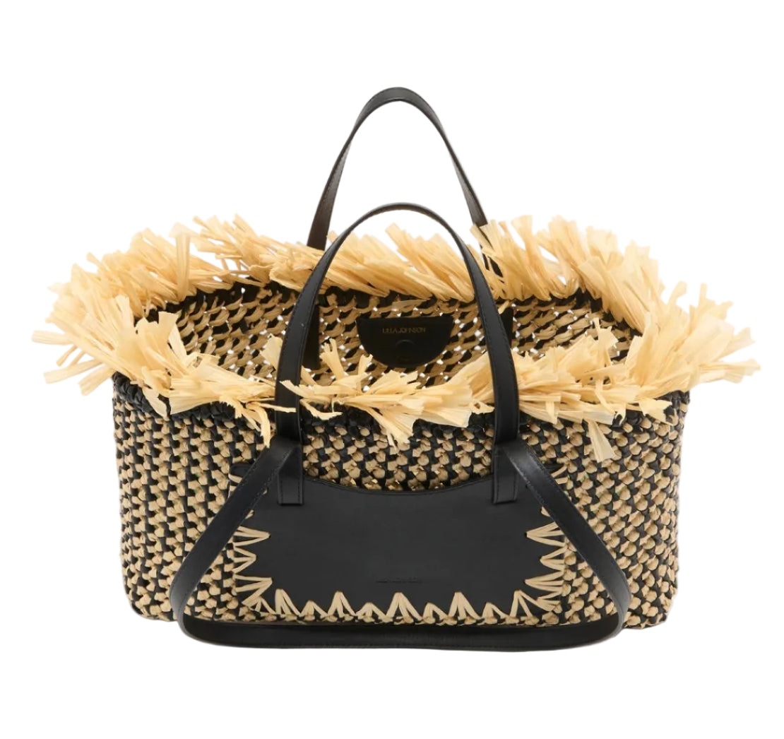 The Lali Small East West Tote by Ulla Johnson is a hand-woven raffia bag with black leather handles and trim, enhanced by beige straw fringe accents along the opening for extra style.